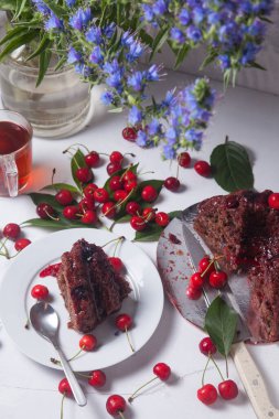 Beyaz arkaplanda kompozisyon - tatlı kirazlı lezzetli çikolatalı pasta, turta parçalı beyaz tabak, bir fincan çay ve bir bardak kavanoz ve güzel bir buket mavi çiçek.