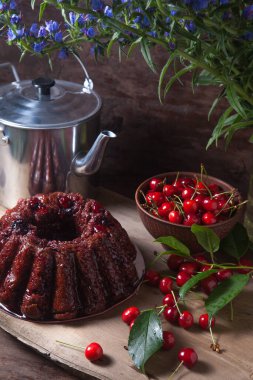 Koyu renk ahşap arka planda kompozisyon - tatlı kirazlı lezzetli çikolatalı pasta, kiraz meyveli kiraz kabı ve kiraz ağacı yaprakları, klasik çaydanlık ve cam kavanoz ve güzel bir buket mavi çiçek.