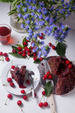 Beyaz arkaplanda kompozisyon - tatlı kirazlı lezzetli çikolatalı pasta, turta parçalı beyaz tabak, bir fincan çay ve bir bardak kavanoz ve güzel bir buket mavi çiçek.