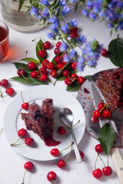 Beyaz arkaplanda kompozisyon - tatlı kirazlı lezzetli çikolatalı pasta, turta parçalı beyaz tabak, bir fincan çay ve bir bardak kavanoz ve güzel bir buket mavi çiçek.
