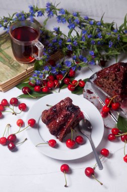 Beyaz arkaplanda kompozisyon - tatlı kirazlı lezzetli çikolatalı pasta, tart parçalı beyaz tabak, klasik kitapta bir fincan çay ve beyaz arka planda mavi çiçeklerin dalı