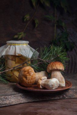 Yenebilir orman mantarları. Üç porcini mantarlı kil levha (sepet, porcino veya kral bolete, genellikle Boletus Edulis olarak bilinir) ve çam ağacı dallı eski ahşap arka planda konserve mantarlı cam kavanoz.