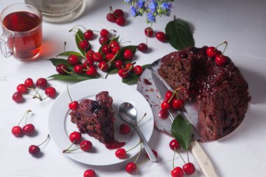 Beyaz arkaplanda kompozisyon - tatlı kirazlı lezzetli çikolatalı pasta, turta parçalı beyaz tabak, bir fincan çay ve bir bardak kavanoz ve güzel bir buket mavi çiçek.