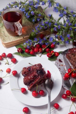 Beyaz arkaplanda kompozisyon - tatlı kirazlı lezzetli çikolatalı pasta, tart parçalı beyaz tabak, klasik kitapta bir fincan çay ve beyaz arka planda mavi çiçeklerin dalı