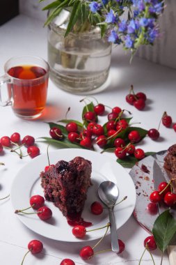 Beyaz arkaplanda kompozisyon - tatlı kirazlı lezzetli çikolatalı pasta, turta parçalı beyaz tabak, bir fincan çay ve bir bardak kavanoz ve güzel bir buket mavi çiçek.