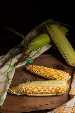 İki tane olgun mısır, yeşil yapraklar ve eski ahşap arka planda yeşil havlu. Beyaz ve sarı taneli koçanlar.