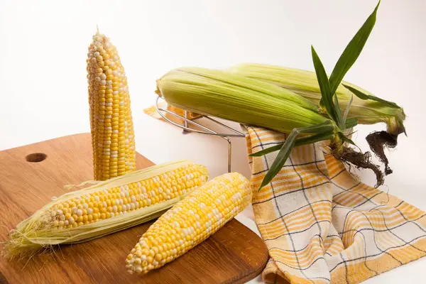Cutting board with three  ears of ripe sweet corn and yellow towel on vintage white wooden background. Cobs with white and yellow grains. Fresh ears of corn with green leaves on background.