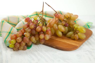 Beyaz üzerine olgun pembe üzümlerle tahta kesmek. Beyaz ahşap zemin üzerinde mavi mutfak havlusu olan pembe şarap üzümleri.