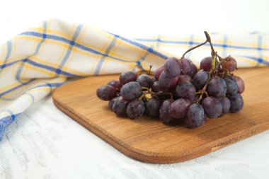 Beyaz üzerine koyu mavi üzümlerden oluşan büyük bir demet kesme tahtası. Mavi mutfak havlusu ve beyaz ahşap arka planda mor şarap üzümleri.