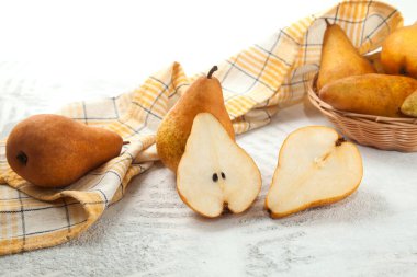 Several whole and halves of ripe juicy pear fruit, wicker basket with pears and yellow kitchen towel on white wooden background