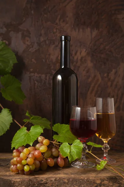 The concept of a delicious alcoholic drink, wine and grapes. Wine bottle, two glasses of red and white wine and bunch of pink grapes on dark rustic wooden background with grapes leaves