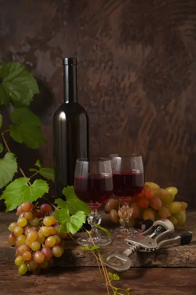 The concept of a delicious alcoholic drink, wine and grapes. Wine bottle, cork, corkscrew, glasses of red wine and bunches of grapes on dark rustic wooden background with grapes leaves