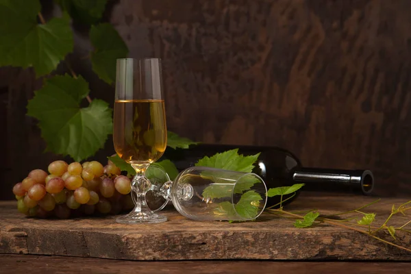 The concept of a delicious alcoholic drink, wine and grapes. Wine bottle, empty glass and glass of white wine and bunch of pink grapes on dark rustic wooden background with grapes leaves