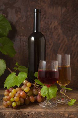 Lezzetli bir alkollü içecek, şarap ve üzüm konsepti. Şarap şişesi, iki bardak kırmızı ve beyaz şarap ve koyu kırsal arka planda üzüm yapraklı bir demet pembe üzüm.