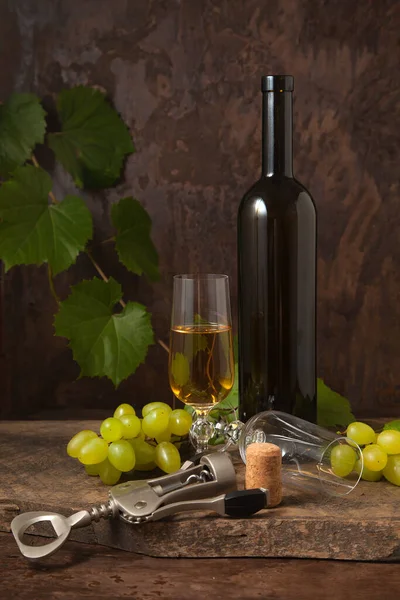 The concept of a delicious alcoholic drink, wine and grapes. Wine bottle, cork, corkscrew, empty glass and glass of white wine and bunch of pink grapes on dark rustic wooden background with grapes leaves