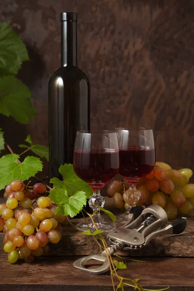 The concept of a delicious alcoholic drink, wine and grapes. Wine bottle, cork, corkscrew, glasses of red wine and bunches of grapes on dark rustic wooden background with grapes leaves