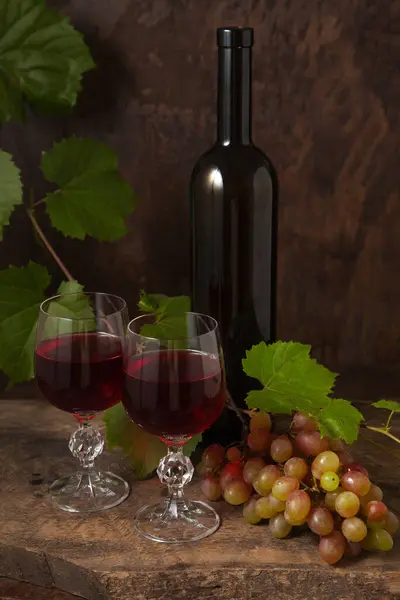 The concept of a delicious alcoholic drink, wine and grapes. Wine bottle, glasses of red wine and bunch of grapes on dark rustic wooden background with grapes leaves