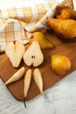 Wooden cutting board with whole, halves and slices of ripe juicy pear fruits, wicker basket with pears and yellow kitchen towel on white wooden background