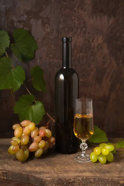 The concept of a delicious alcoholic drink, wine and grapes. Wine bottle, glass of white wine and bunches of pink and green grapes  on dark rustic wooden background with grapes leaves