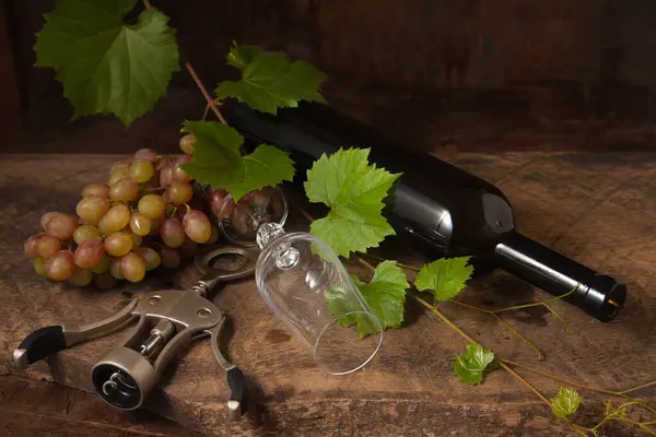 The concept of a delicious alcoholic drink, wine and grapes. Top view of wine bottle, corkscrew, empty wine glass and bunch of pink grapes on dark rustic wooden background with grapes leaves