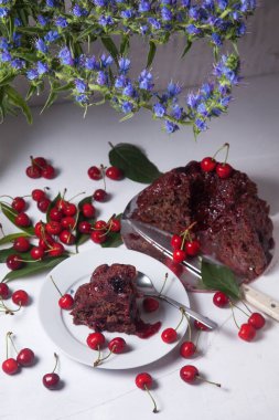 Beyaz arkaplanda kompozisyon - tatlı kirazlı lezzetli çikolatalı pasta, turta parçalı beyaz tabak, bir fincan çay ve bir bardak kavanoz ve güzel bir buket mavi çiçek.