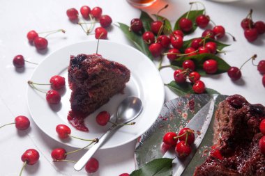 Beyaz arkaplanda kompozisyon - tatlı kirazlı lezzetli çikolatalı pasta, turta parçalı beyaz tabak, bir fincan çay ve bir bardak kavanoz ve güzel bir buket mavi çiçek.
