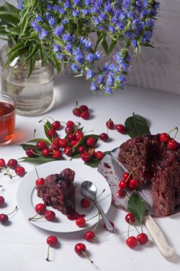 Beyaz arkaplanda kompozisyon - tatlı kirazlı lezzetli çikolatalı pasta, turta parçalı beyaz tabak, bir fincan çay ve bir bardak kavanoz ve güzel bir buket mavi çiçek.