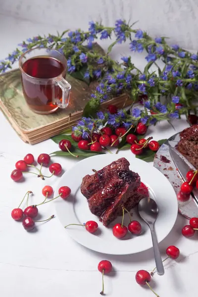 Beyaz arkaplanda kompozisyon - tatlı kirazlı lezzetli çikolatalı pasta, tart parçalı beyaz tabak, klasik kitapta bir fincan çay ve beyaz arka planda mavi çiçeklerin dalı