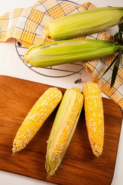 Cutting board with three  ears of ripe sweet corn and yellow towel on vintage white wooden background. Cobs with white and yellow grains. Fresh ears of corn with green leaves on background.