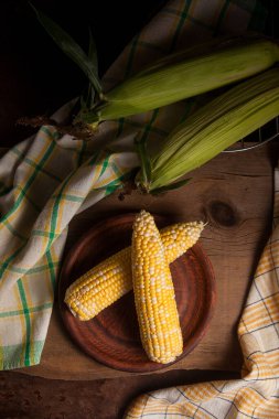 Kaliteli ahşap arka planda olgun mısır ve yeşil havluyla iki kulaklı kil tabağı. Beyaz ve sarı taneli koçanlar. Arka planda yeşil yapraklı taze mısır kulakları.