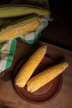 Kaliteli ahşap arka planda olgun mısır ve yeşil havluyla iki kulaklı kil tabağı. Beyaz ve sarı taneli koçanlar. Arka planda yeşil yapraklı taze mısır kulakları.