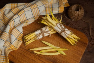 Tahta kesme tahtası, iki deste ve ahşap arka planda bir sürü çiğ sarı kapsül ve sarı mutfak havlusu. Fransız fasulyesi ya da Hindistan 'dan String fasulyesi olarak da bilinir. Sebze koleksiyonu.