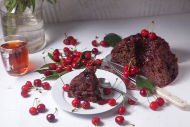 Beyaz arkaplanda kompozisyon - tatlı kirazlı lezzetli çikolatalı pasta, turta parçalı beyaz tabak, bir fincan çay ve bir bardak kavanoz ve güzel bir buket mavi çiçek.