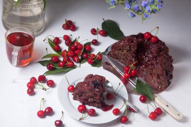 Beyaz arkaplanda kompozisyon - tatlı kirazlı lezzetli çikolatalı pasta, turta parçalı beyaz tabak, bir fincan çay ve bir bardak kavanoz ve güzel bir buket mavi çiçek.