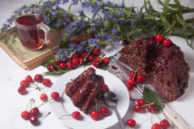 Beyaz arkaplanda kompozisyon - tatlı kirazlı lezzetli çikolatalı pasta, tart parçalı beyaz tabak, klasik kitapta bir fincan çay ve beyaz arka planda mavi çiçeklerin dalı