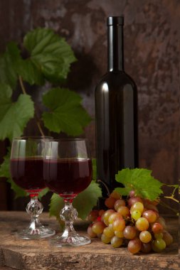 Lezzetli bir alkollü içecek, şarap ve üzüm konsepti. Şarap şişesi, kırmızı şarap bardakları ve koyu kırsal ahşap arka planda üzüm yapraklı üzümler.