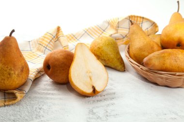 Several whole and halves of ripe juicy pear fruit, wicker basket with pears and yellow kitchen towel on white wooden background