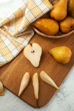 Wooden cutting board with whole, halves and slices of ripe juicy pear fruits, wicker basket with pears and yellow kitchen towel on white wooden background