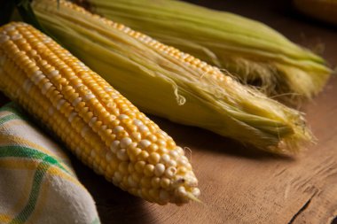 Üç tane olgun, yeşil yapraklı ve eski ahşap arka planda yeşil havluyla tatlı mısırın görüntüsünü kapat. Beyaz ve sarı taneli koçanlar.