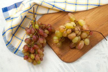 Beyaz üzerine olgun pembe üzümlerle tahta kesmek. Beyaz ahşap zemin üzerinde mavi mutfak havlusu olan pembe şarap üzümleri.