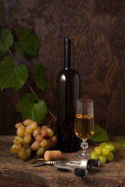 Lezzetli bir alkollü içecek, şarap ve üzüm konsepti. Şarap şişesi, mantar, tirbuşon, bir bardak beyaz şarap ve koyu kırsal arka planda üzümlü pembe ve yeşil üzümler.