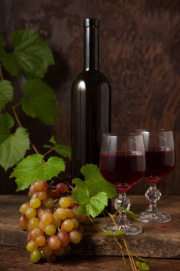 Lezzetli bir alkollü içecek, şarap ve üzüm konsepti. Şarap şişesi, kırmızı şarap bardakları ve koyu kırsal ahşap arka planda üzüm yapraklı üzümler.