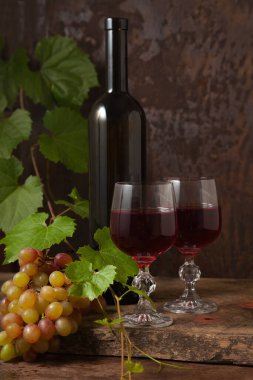 Lezzetli bir alkollü içecek, şarap ve üzüm konsepti. Şarap şişesi, kırmızı şarap bardakları ve koyu kırsal ahşap arka planda üzüm yapraklı üzümler.