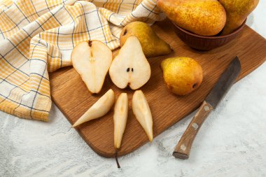 Wooden cutting board with whole, halves and slices of ripe juicy pear fruits, wicker basket with pears and yellow kitchen towel on white wooden background