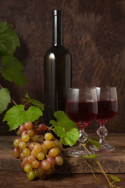 Lezzetli bir alkollü içecek, şarap ve üzüm konsepti. Şarap şişesi, kırmızı şarap bardakları ve koyu kırsal ahşap arka planda üzüm yapraklı üzümler.