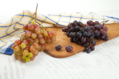 Beyaz üzerine olgun pembe ve mor üzümlerden oluşan büyük bir demet kesme tahtası. Beyaz ahşap arka planda mavi mutfak havlusu olan pembe ve mavi şarap üzümleri.