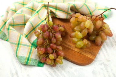 Beyaz üzerine olgun pembe üzümlerle tahta kesmek. Beyaz ahşap zemin üzerinde mavi mutfak havlusu olan pembe şarap üzümleri.