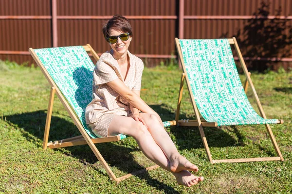 Young woman in pajama is resting in chair on a green lawn on sunny summer day - village and country life