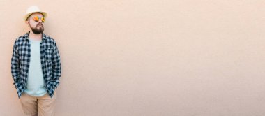 Yazlık şapka ve ekose gömlek giyen yakışıklı beyaz adam duvarın yanında mutlu bir şekilde gülümsüyor.