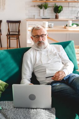 Üst düzey yaşlı adam dizüstü bilgisayarla online ödeme yapıyor. Gülümseyen yaşlı büyükbaba kredi kartıyla alışveriş yapıyor. Emeklinin elinde internet bankacılığı için banka kartı var